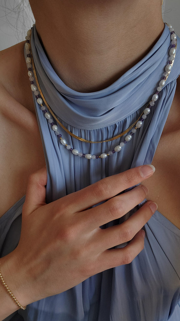 Photo of a woman in a light blue collared dress, adjusting the straps to accentuate her neckline. She is wearing two handcrafted necklaces from Blue Trimble Designs: the first is a 14-karat gold-filled herringbone chain, and the second, a beaded necklace adorned with freshwater pearls, amethyst, and rose quartz. Each piece complements the subtle elegance of her outfit.