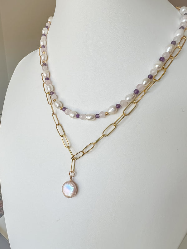 Photo shows a white display bust featuring two elegant necklaces. The first necklace is handcrafted with beads made of freshwater pearls, amethyst, and rose quartz. The second necklace features a round coin pearl drop pendant on a elongated oval chain. Both necklaces are delicately arranged to highlight their unique beauty and craftsmanship