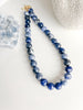 Blue sodalite necklace made with 12 mm round beads, styled in a still life with a crystal and the base of a vase on a white background.