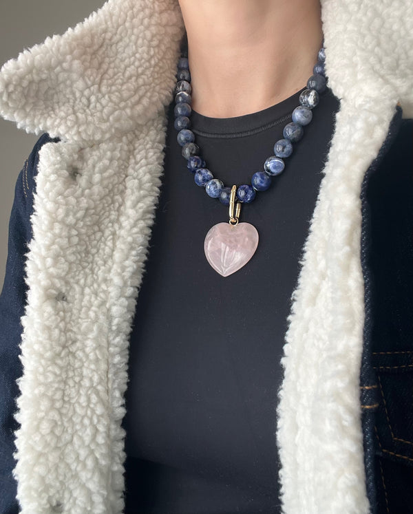 Blue sodalite beaded necklace with a polished 35 mm rose quartz heart on a carabiner push clasp, worn by a model in a black T-shirt and a denim shearling-collared coat.