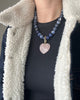 Blue sodalite beaded necklace with a polished 35 mm rose quartz heart on a carabiner push clasp, worn by a model in a black T-shirt and a denim shearling-collared coat.