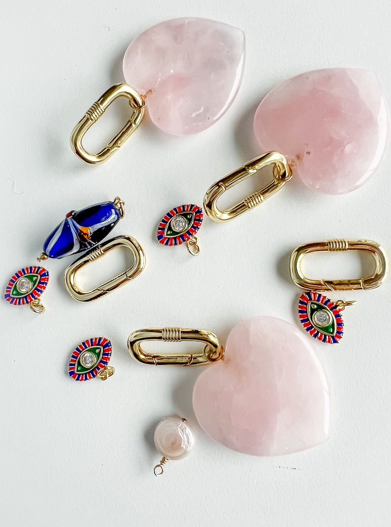 Charm Garden collection featuring 35 mm rose quartz heart charms, red, blue, and green evil eye pendants with cubic zirconia centers, gold carabiner push clasps, a freshwater pearl, and a blue glass bead, arranged on a white background.