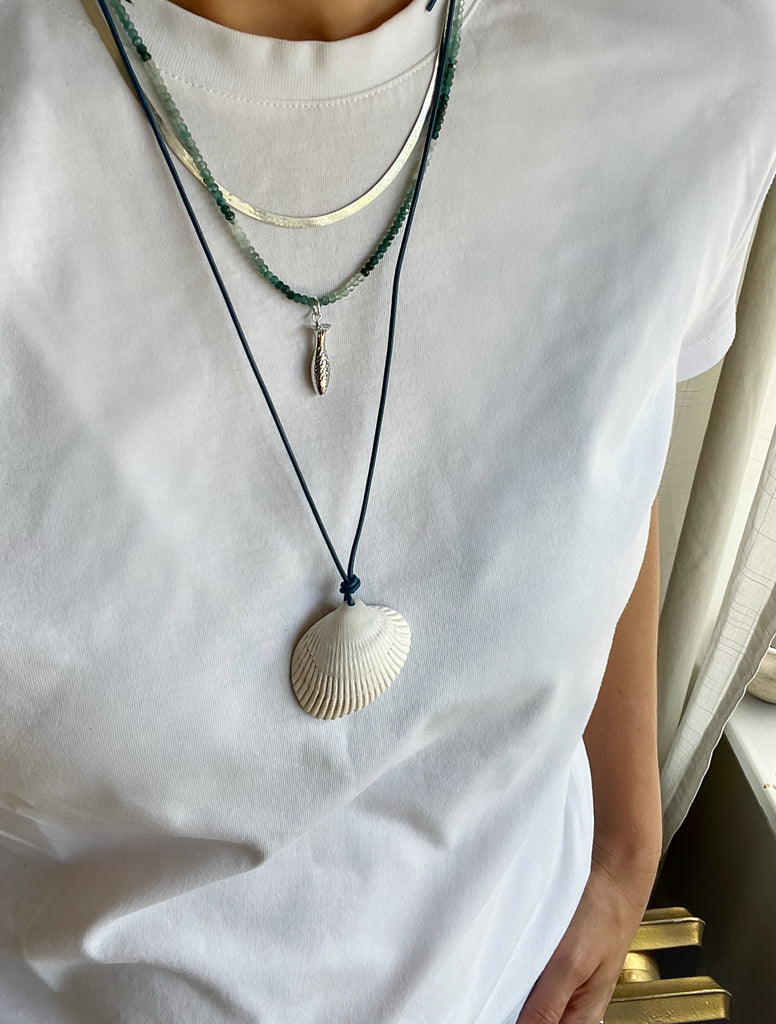 Green grandidierite crystal necklace with a sterling silver fish charm layered with a leather shell necklace and a sterling silver herringbone chain over a white T-shirt.