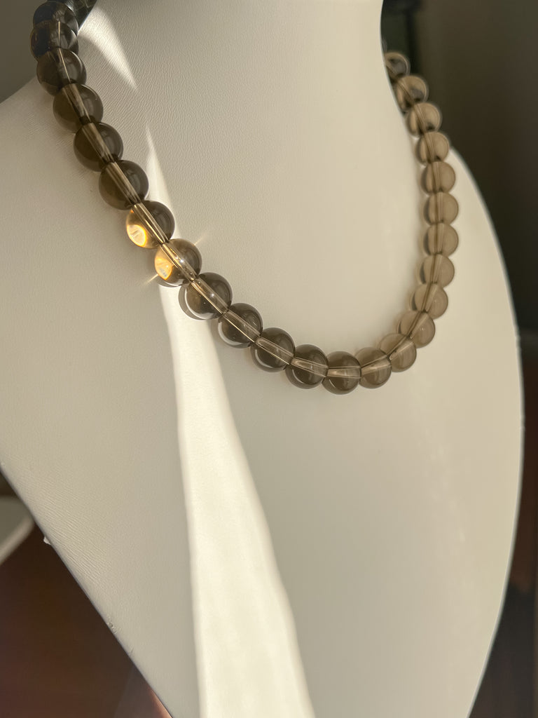 12 mm round smoky quartz strand necklace displayed on a white jewelry bust with sunlight highlighting the beads.