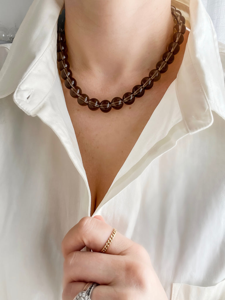 12 mm round smoky quartz strand necklace worn by a model over a white button-up shirt, showing how it sits naturally while she holds the buttons.